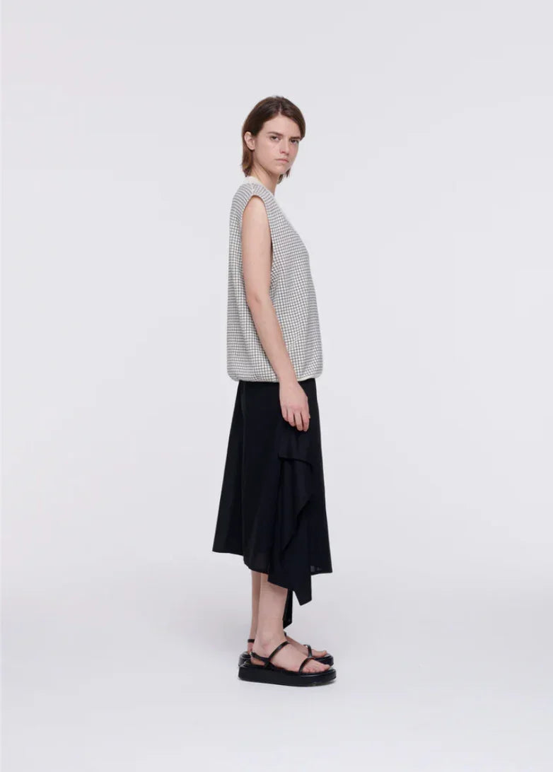 Side view of woman wearing sleeveless checkered top, black asymmetrical skirt, and black sandals on white background