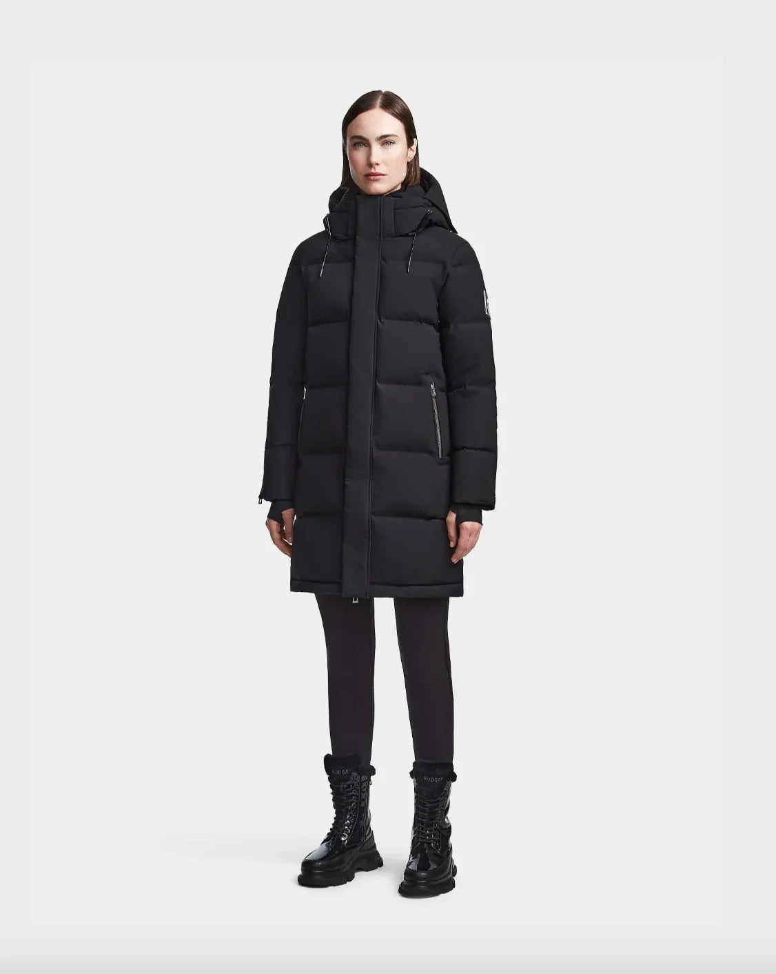 Woman wearing black long down puffer coat with hood, black leggings, and black lace-up boots
