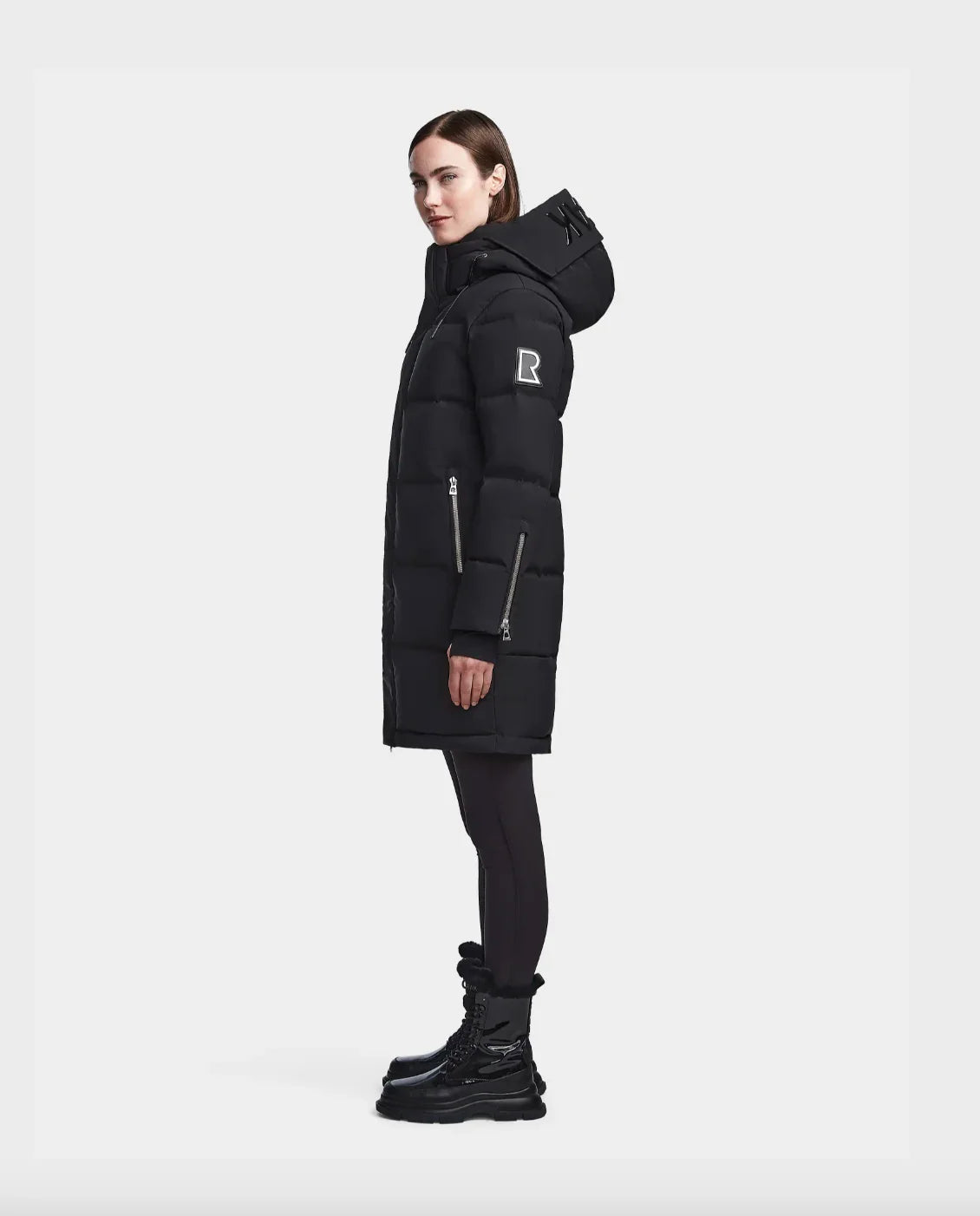 Woman wearing black knee-length puffer winter coat with hood and zipper details, black leggings, and black boots