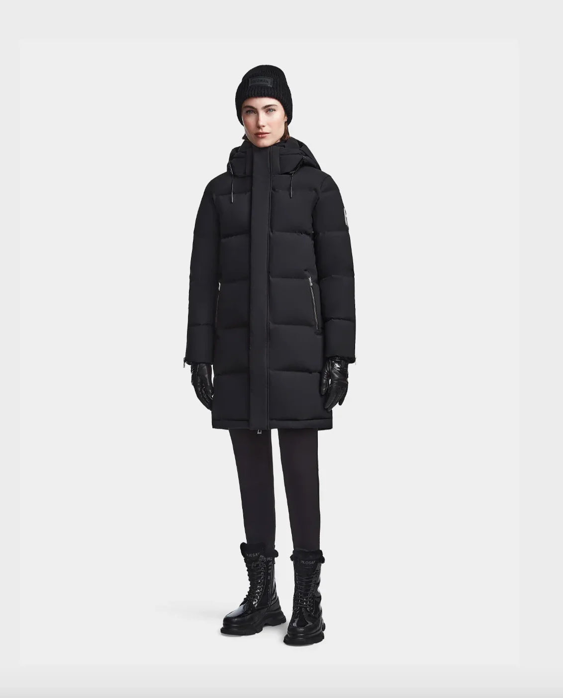 Woman wearing black puffer coat, knit beanie, leather gloves, black leggings, and lace-up boots