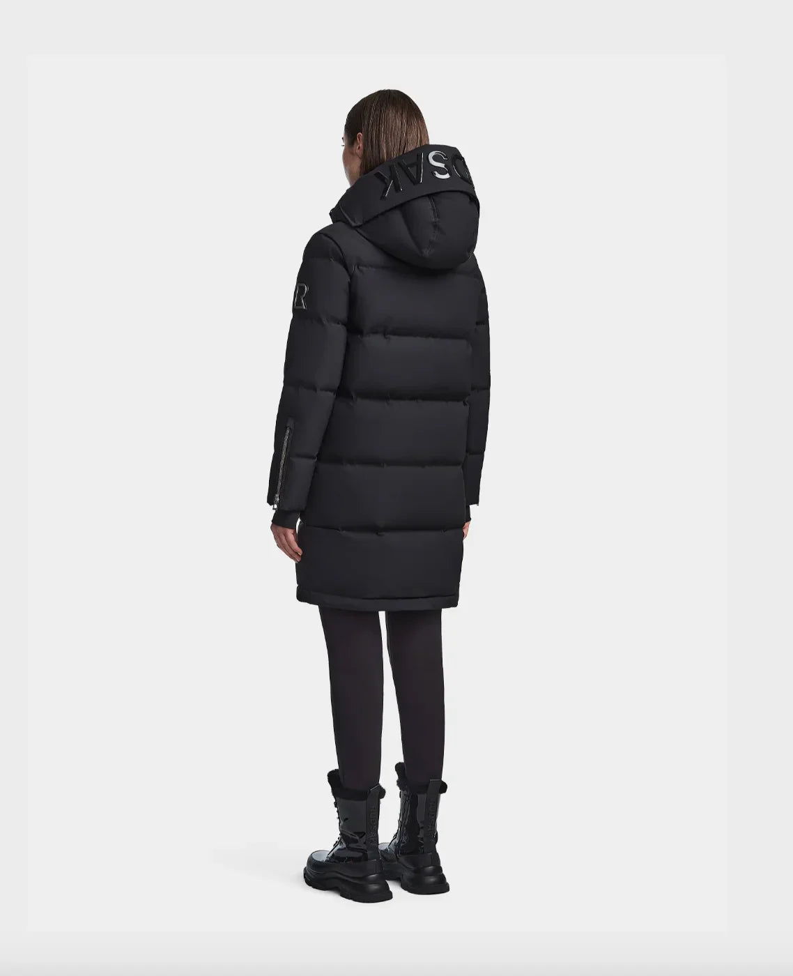 Rear view of woman wearing black puffer coat with hood and black leggings and boots on white background