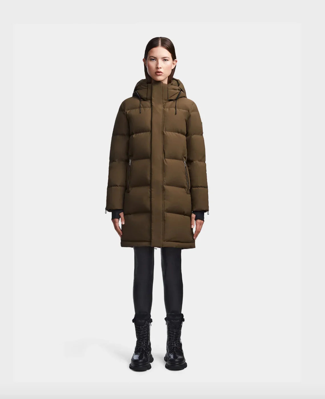 Woman wearing olive green long puffer winter coat with hood and black boots on white background