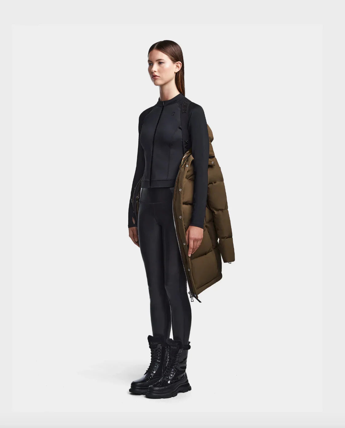 Woman in black zip-up sportswear and leggings holding an olive green puffer jacket with black lace-up boots