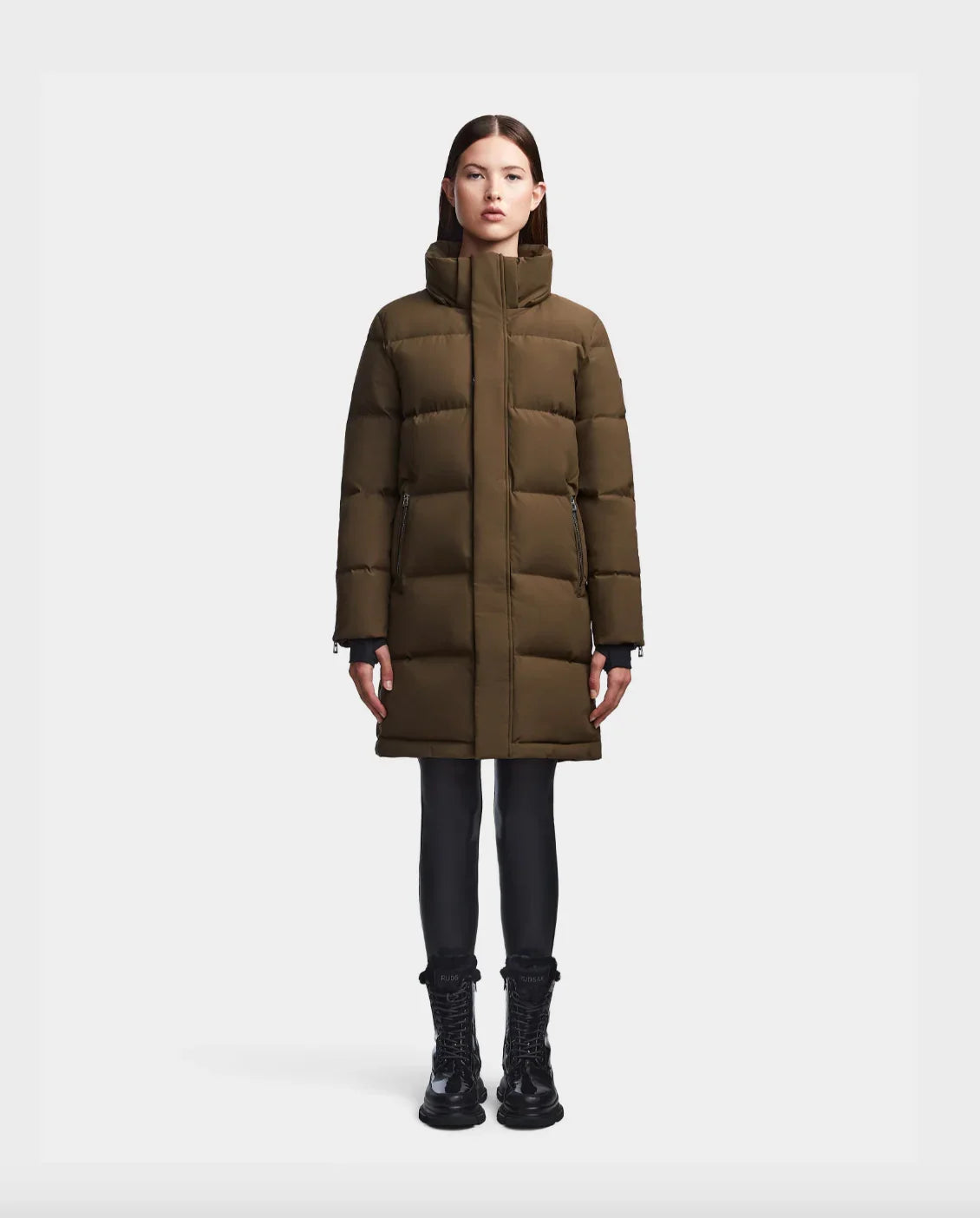 Woman wearing long brown puffer coat with high collar and black boots on white background