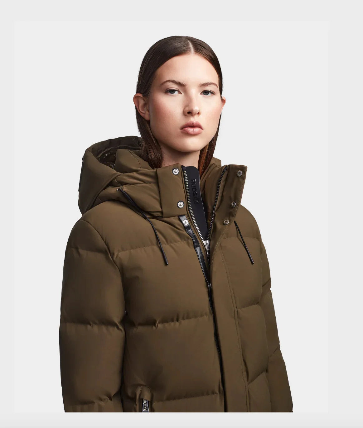 Woman wearing olive green puffer jacket with hood and black zipper details on white background