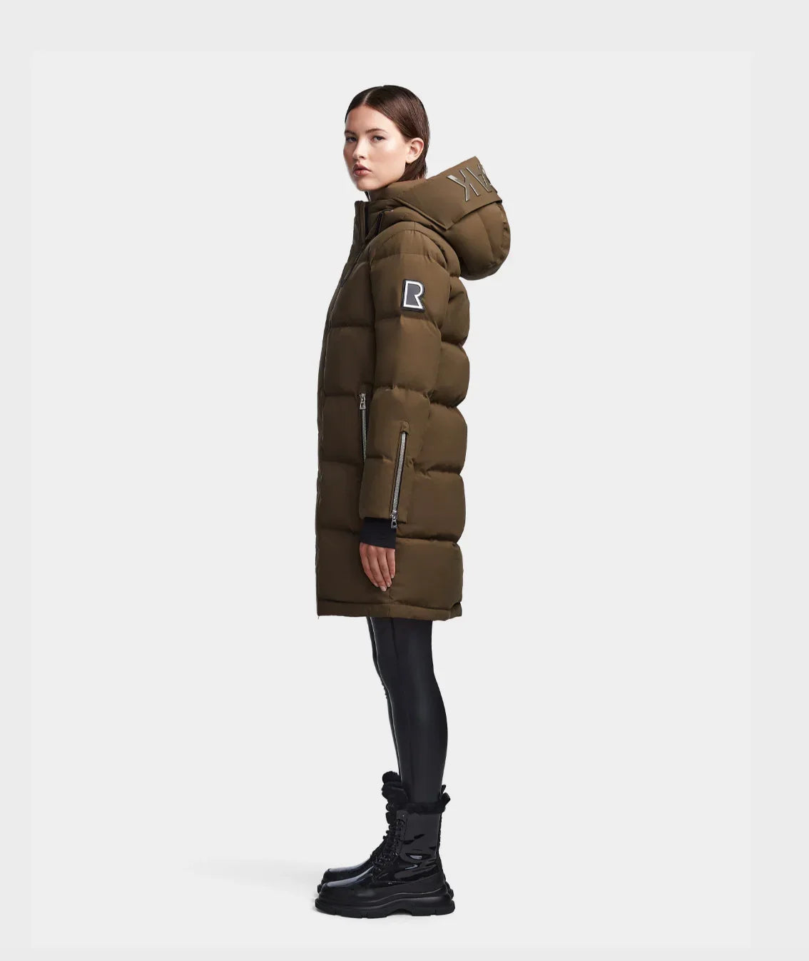 Side view of woman wearing olive green long puffer coat with hood and black boots on white background