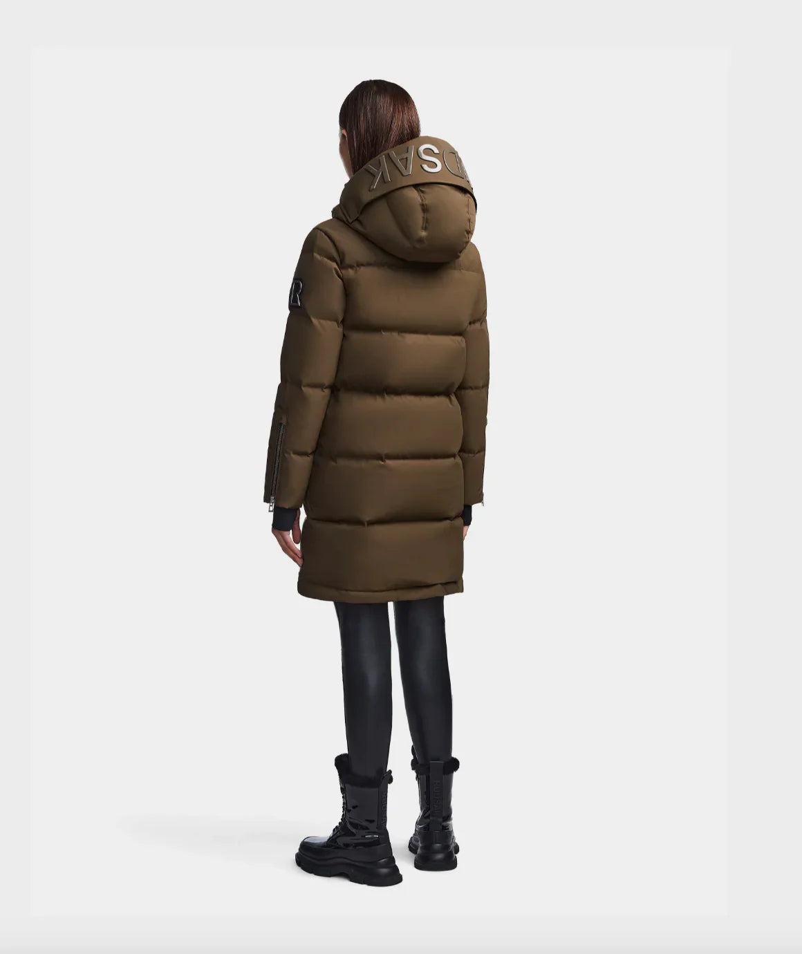 Woman wearing olive green padded long winter coat with hood and black boots, back view