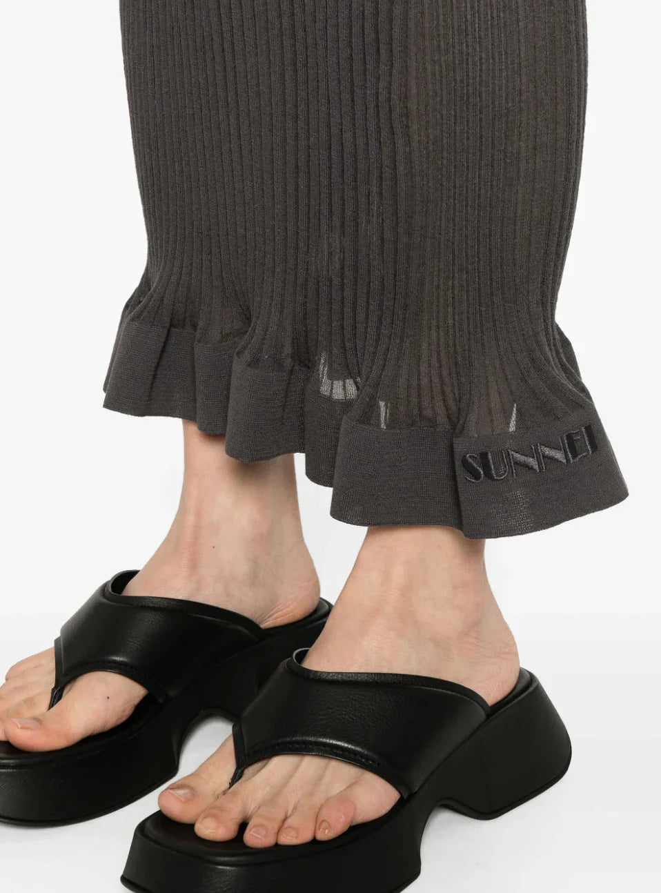 Close-up of black ribbed pants with frill hem and 'SUNNEI' embroidery worn with black platform sandals