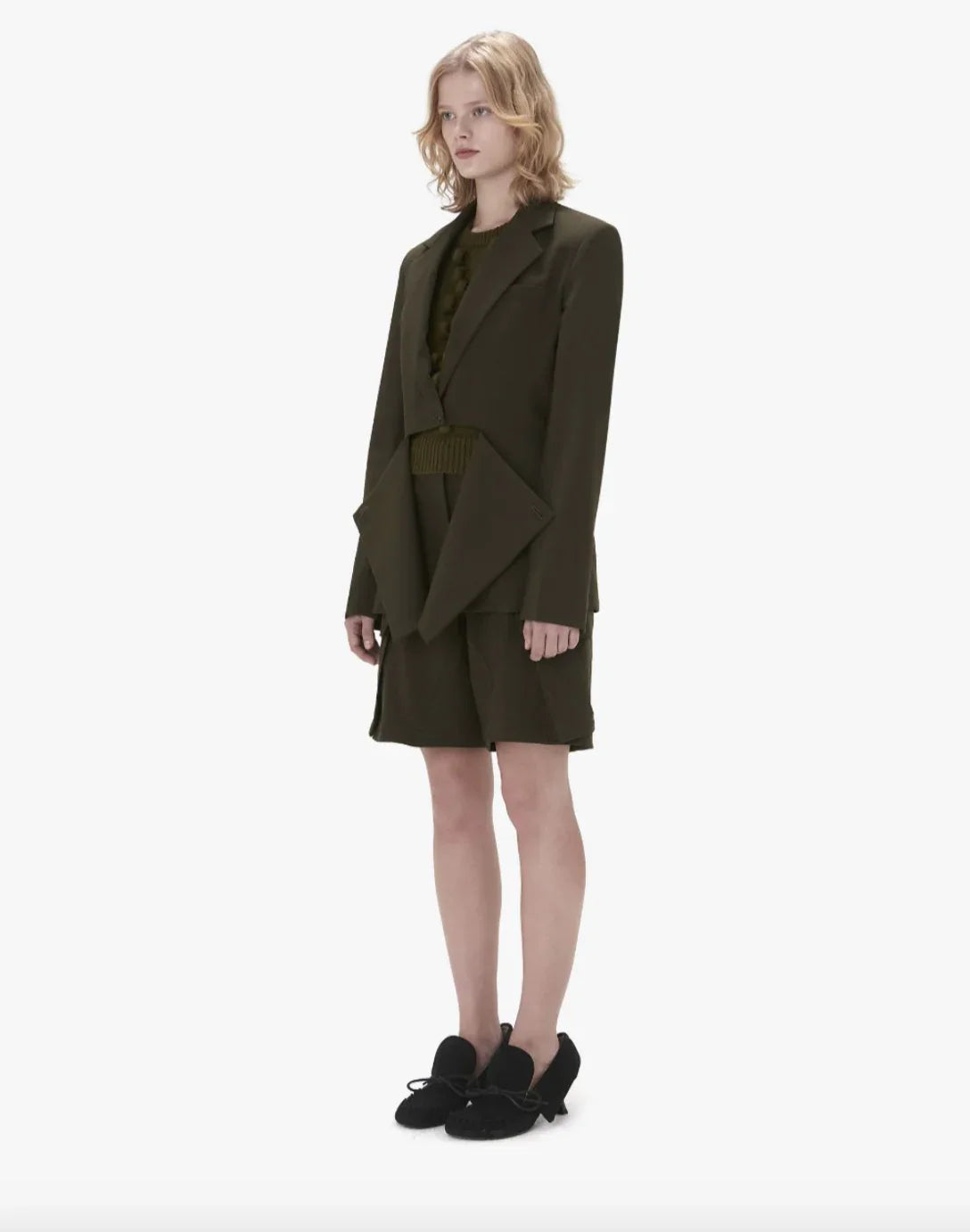 Model wearing a dark olive asymmetrical blazer, matching shorts, sweater, and black lace-up shoes
