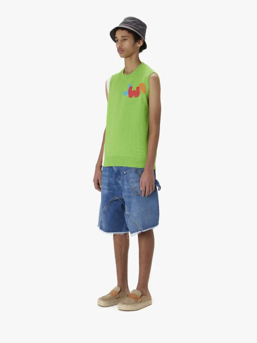Male model wearing lime green sleeveless knit top with colorful logo, denim shorts, gray bucket hat, and beige slip-on shoes