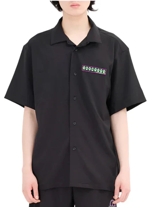 Person wearing black short-sleeve button-up shirt with small green and purple graphic on chest pocket area