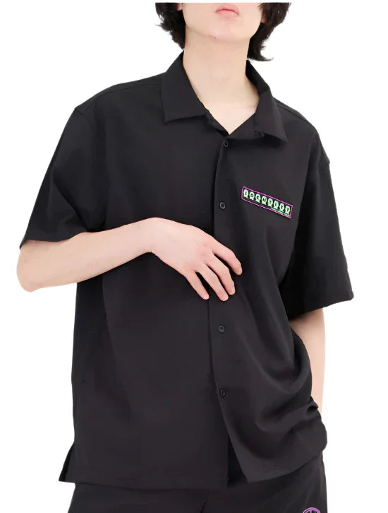 Person wearing black short-sleeve button-up shirt with green and purple logo patch on chest