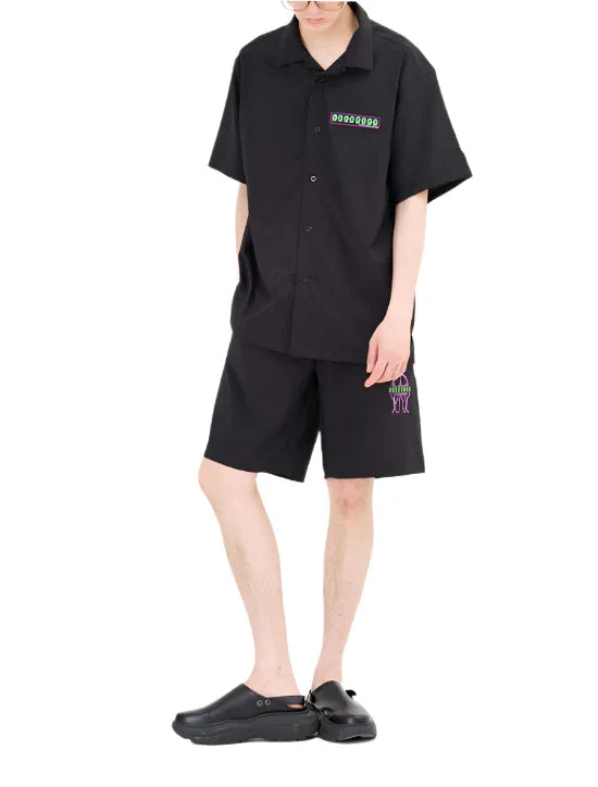 Person wearing black short-sleeve button-up shirt and matching shorts with neon green and purple graphic accents