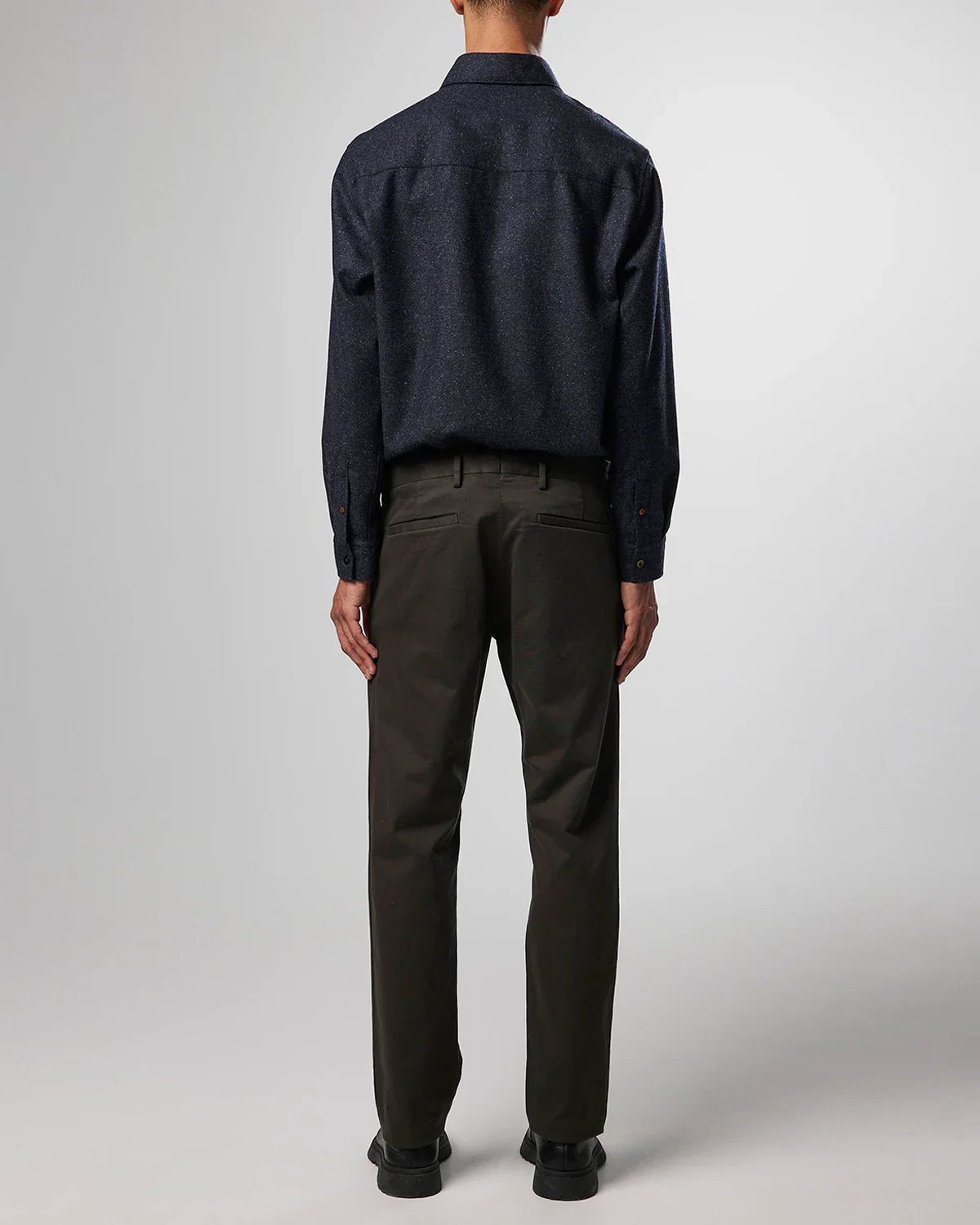 Rear view of man wearing black speckled shirt, dark olive chinos, and black shoes on gray background
