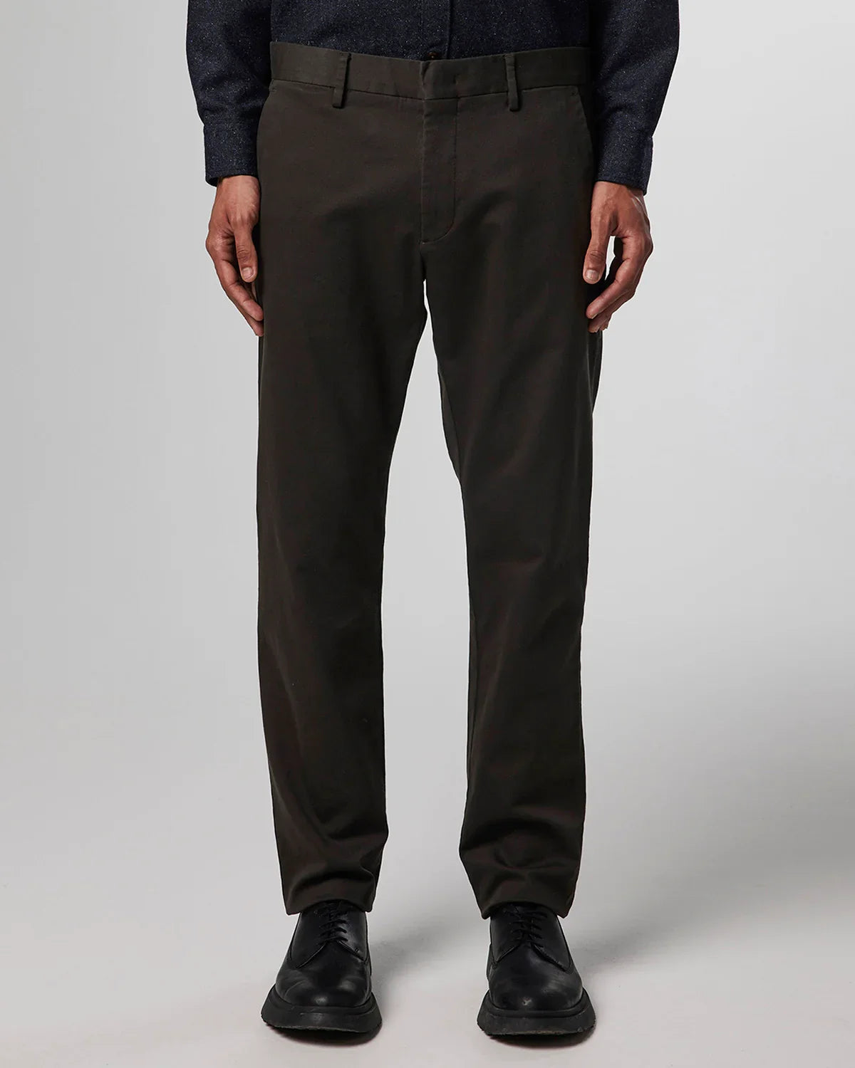 Men's dark brown casual pants worn with black dress shoes and a dark blue shirt on a gray background